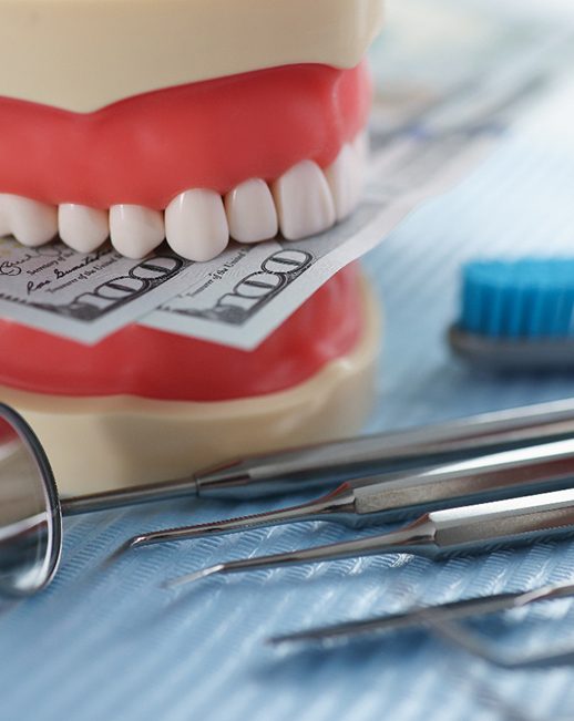 A jaw mockup biting hundred-dollar bills next to some dental tools
